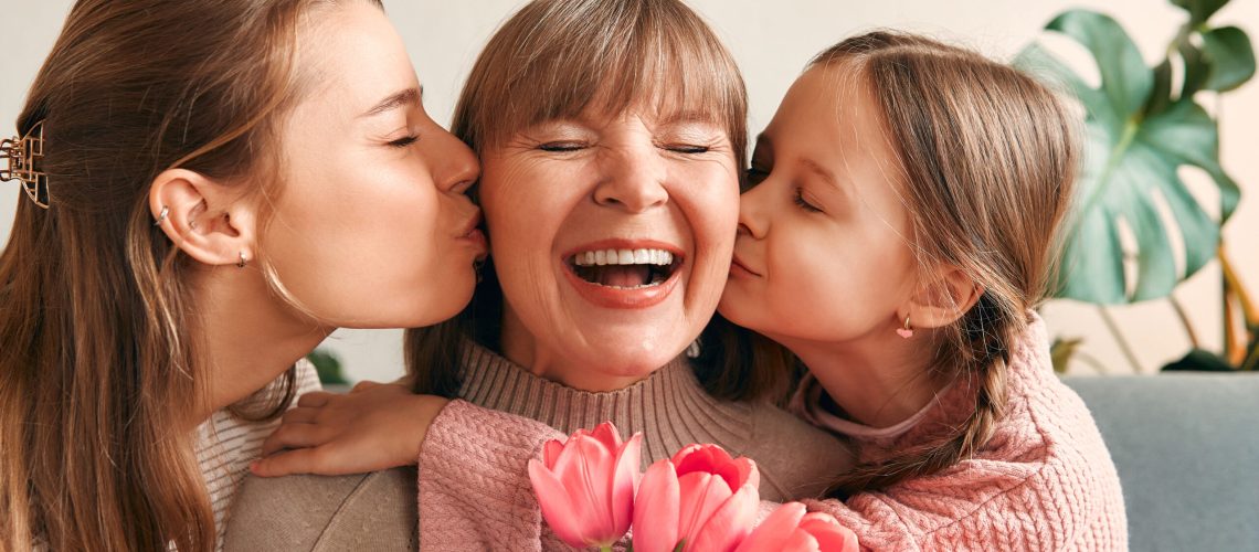 Daughter and granddaughter kissing grandmother in the living room of the house and giving her flowers. Concept of Women's Day and Mother's Day. Women's generation.