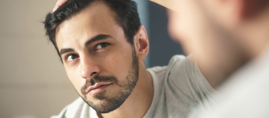 Latino person with beard grooming in bathroom at home. White metrosexual man worried for hair loss and looking at mirror his receding hairline.